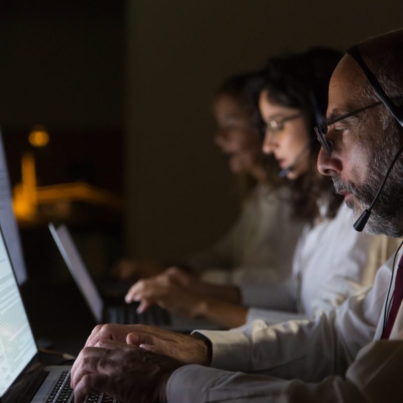 Focused coworkers in headsets typing on laptops. Side view of focused call center operators in headsets using laptop computers in dark office. Working late concept